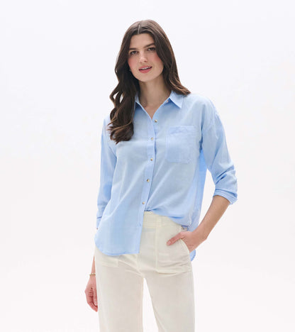 Light blue chambray button-down shirt with collar, chest pocket, and long sleeves, worn with cream pants.