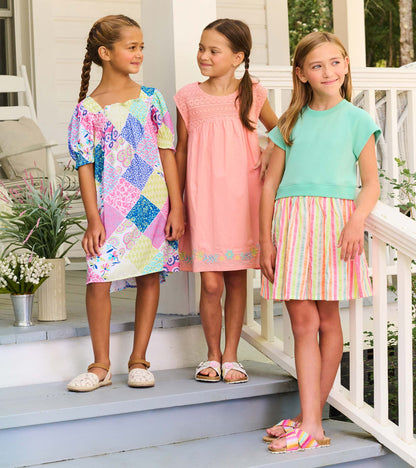 Three girls wearing colorful summer dresses, including patchwork print, pink, and mint with striped skirt, standing on porch…