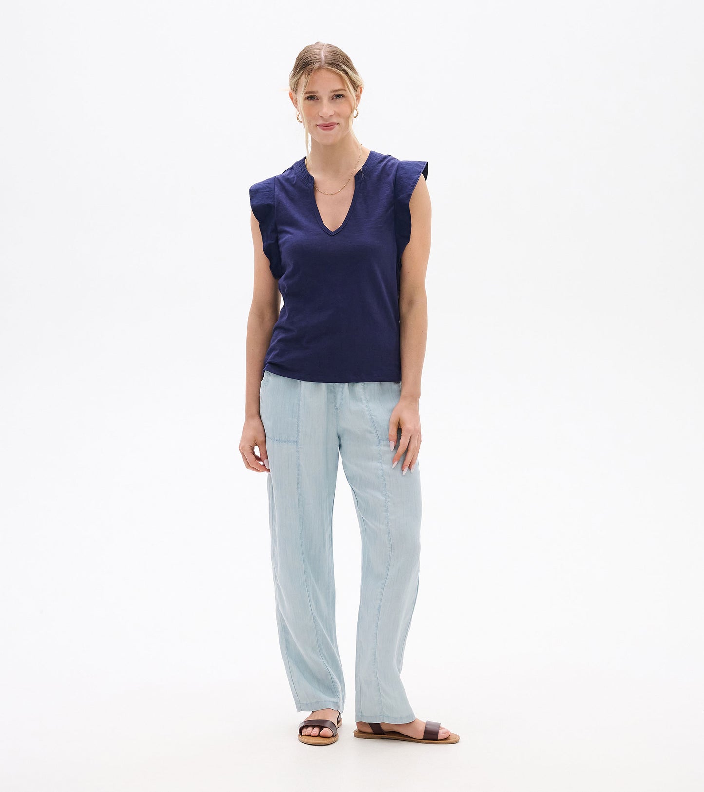 Woman wearing a navy sleeveless V-neck tank top with ruffled shoulders and light blue pants, front view