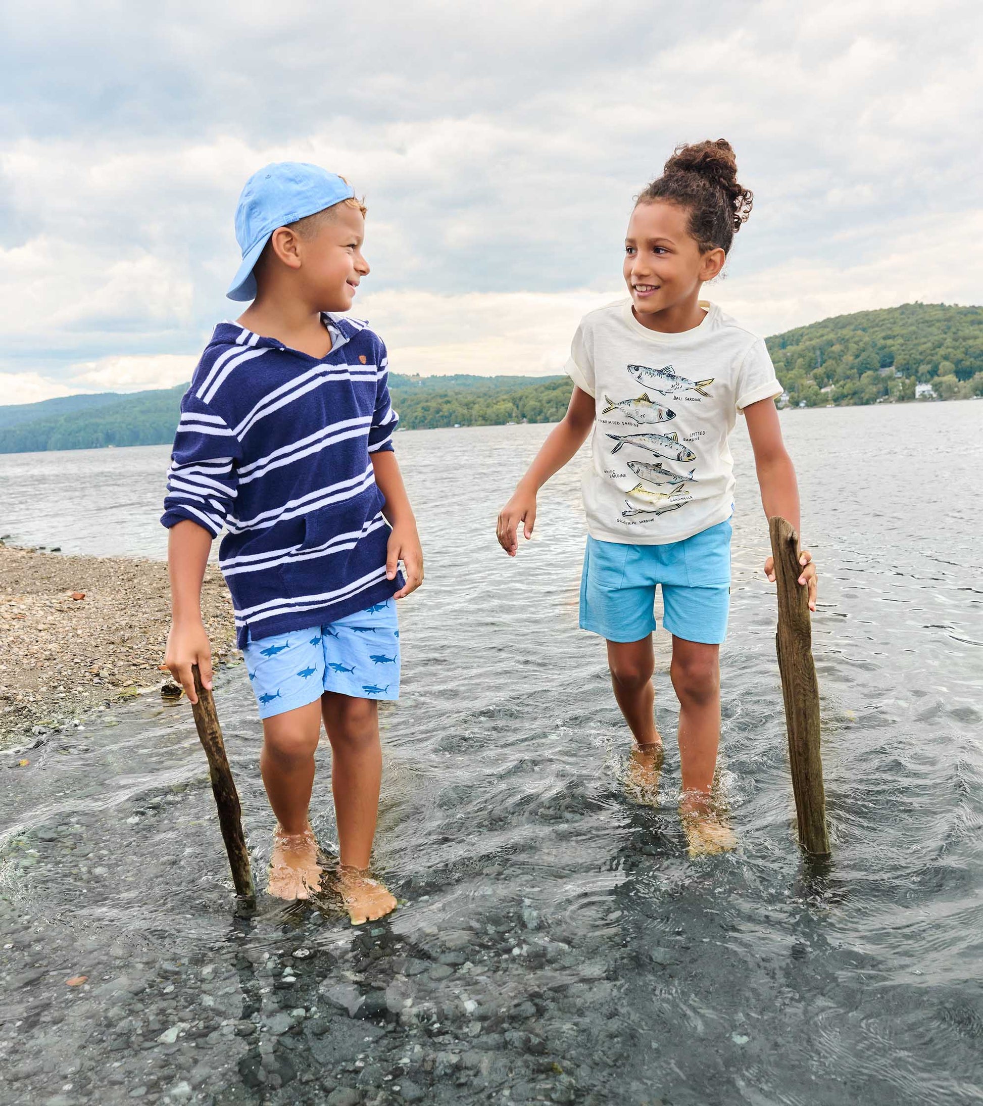 Two children wearing graphic tees and shorts standing in shallow water by a lake