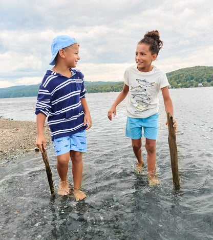 Two children wearing graphic tees and shorts standing in shallow water by a lake