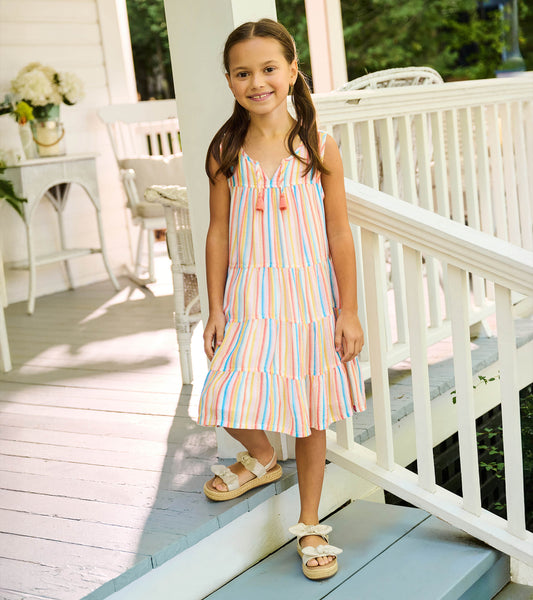 Girl wearing a sleeveless pastel striped tiered dress with tassel ties, shown full length on porch steps