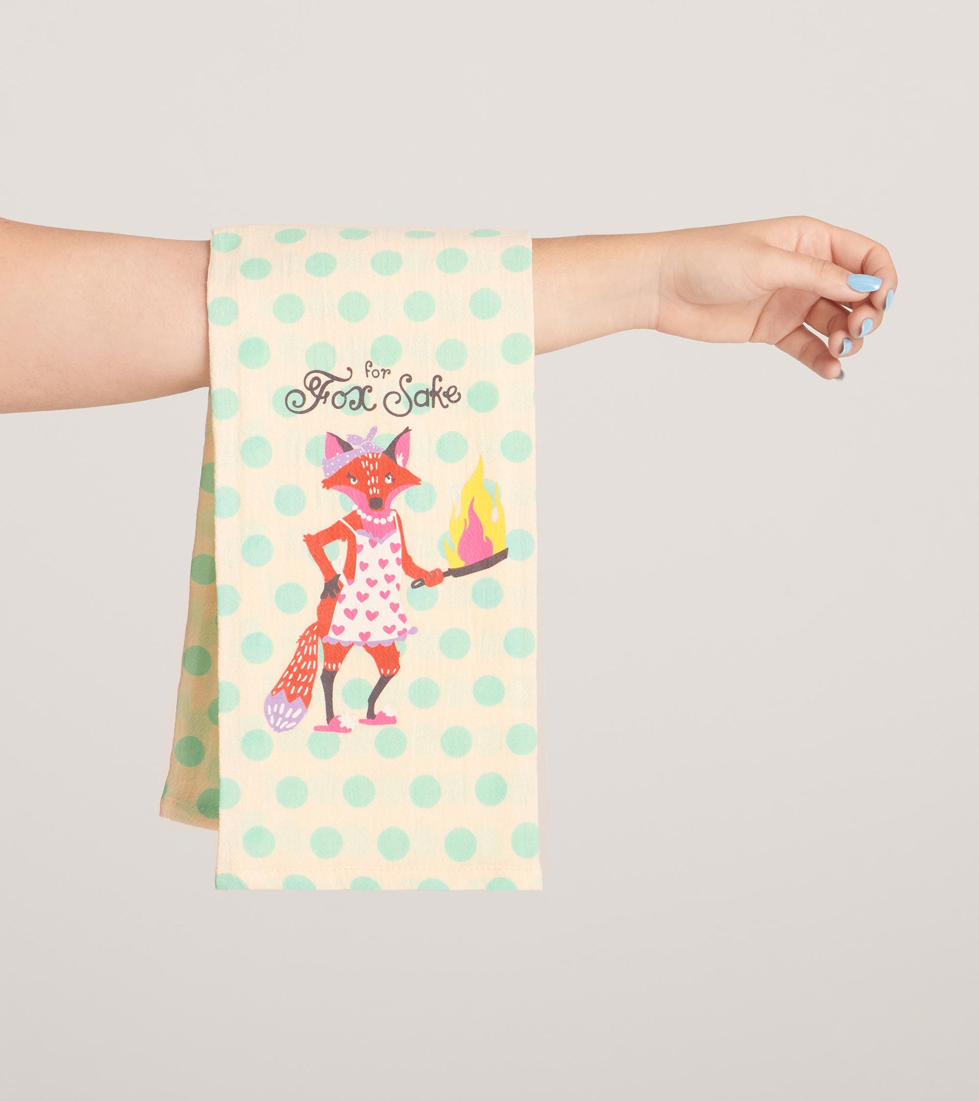 Cream tea towel with mint polka dots and a whimsical fox character holding a flaming pan, perfect for kitchen use.