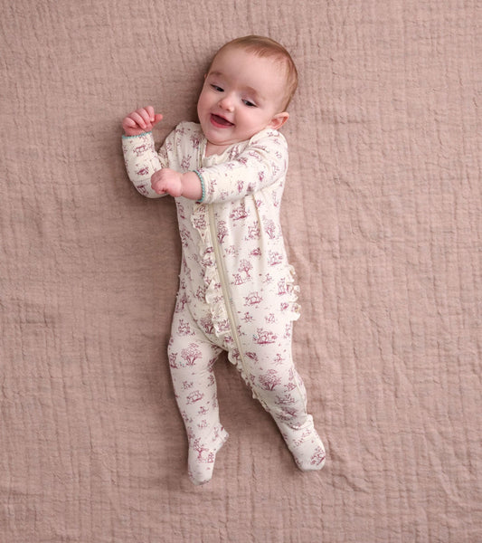 Footed sleeper in cream with pink animal print and ruffle details, worn by a smiling baby on a soft, textured surface.