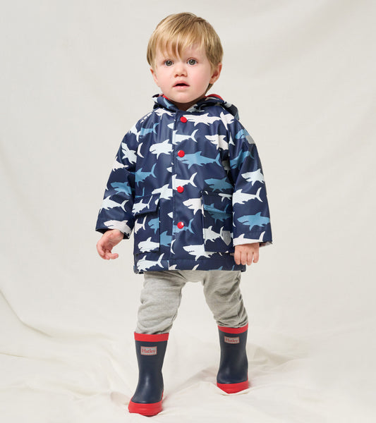 Navy baby raincoat with shark print, red buttons, and hood, worn over gray pants and rain boots.