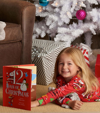 Red pajama set with festive patterns, featuring a child reading a Christmas book by a decorated tree and wrapped gifts.