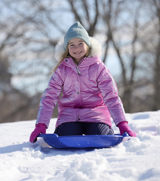 Pink puffer jacket with a shiny finish, worn by a girl sitting on a sled in the snow, paired with a light blue beanie.