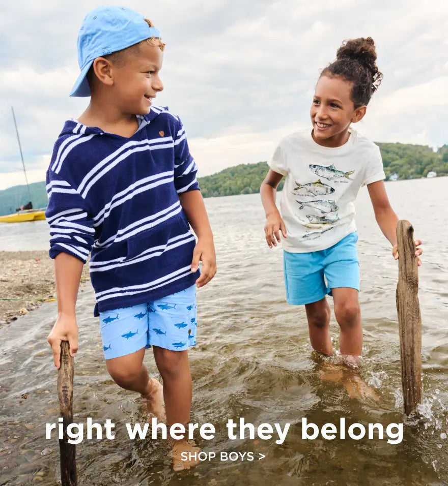 Shop boys' clothing. Showing two kids wading at a lakeshore in casual summer outfits.