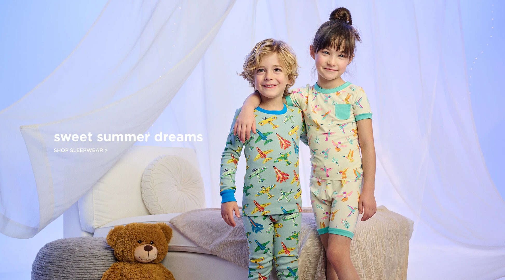 Promotes kids' sleepwear collection. Showing two children wearing patterned pajamas beside a bed and teddy bear.